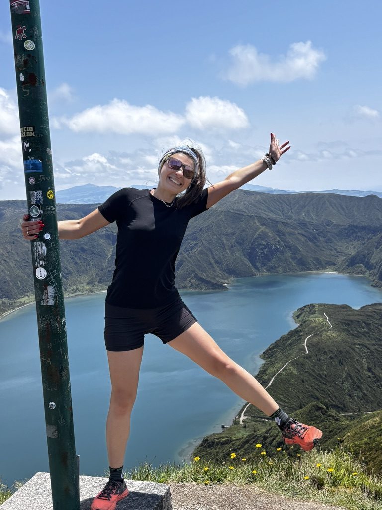 Anca with arms outstretched on a hilltop summit above a volcanic crater lake in the Azores, pure exhilaration