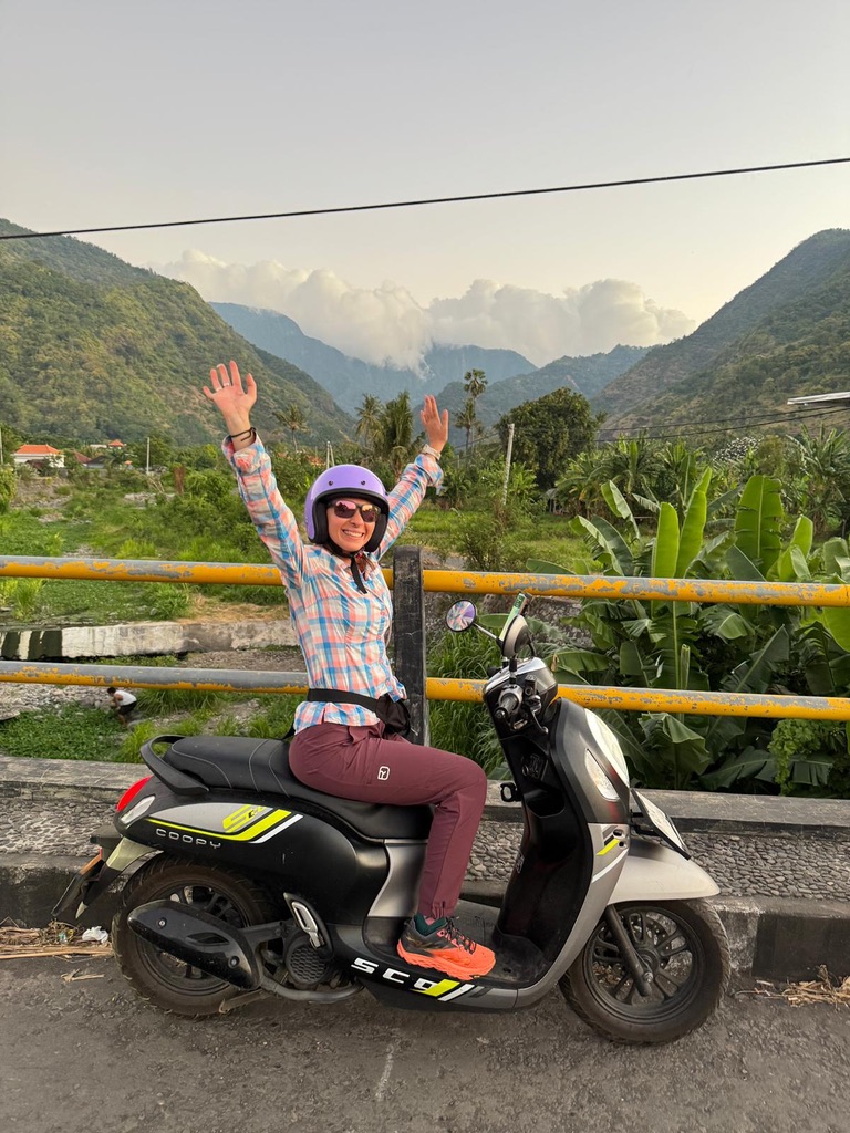Anca on a scooter in Indonesia, arms raised, green mountains behind